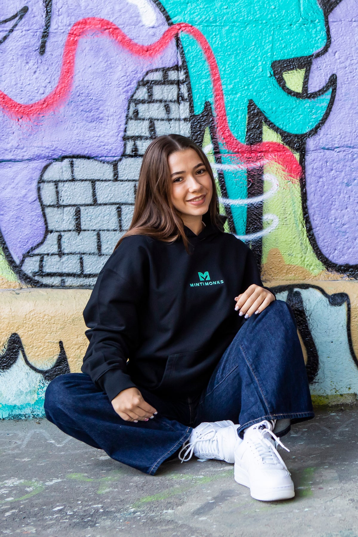 Schwarzer Mintimonks-Hoodie mit gesticktem Logo, getragen vor einer Graffitiwand in Wien
